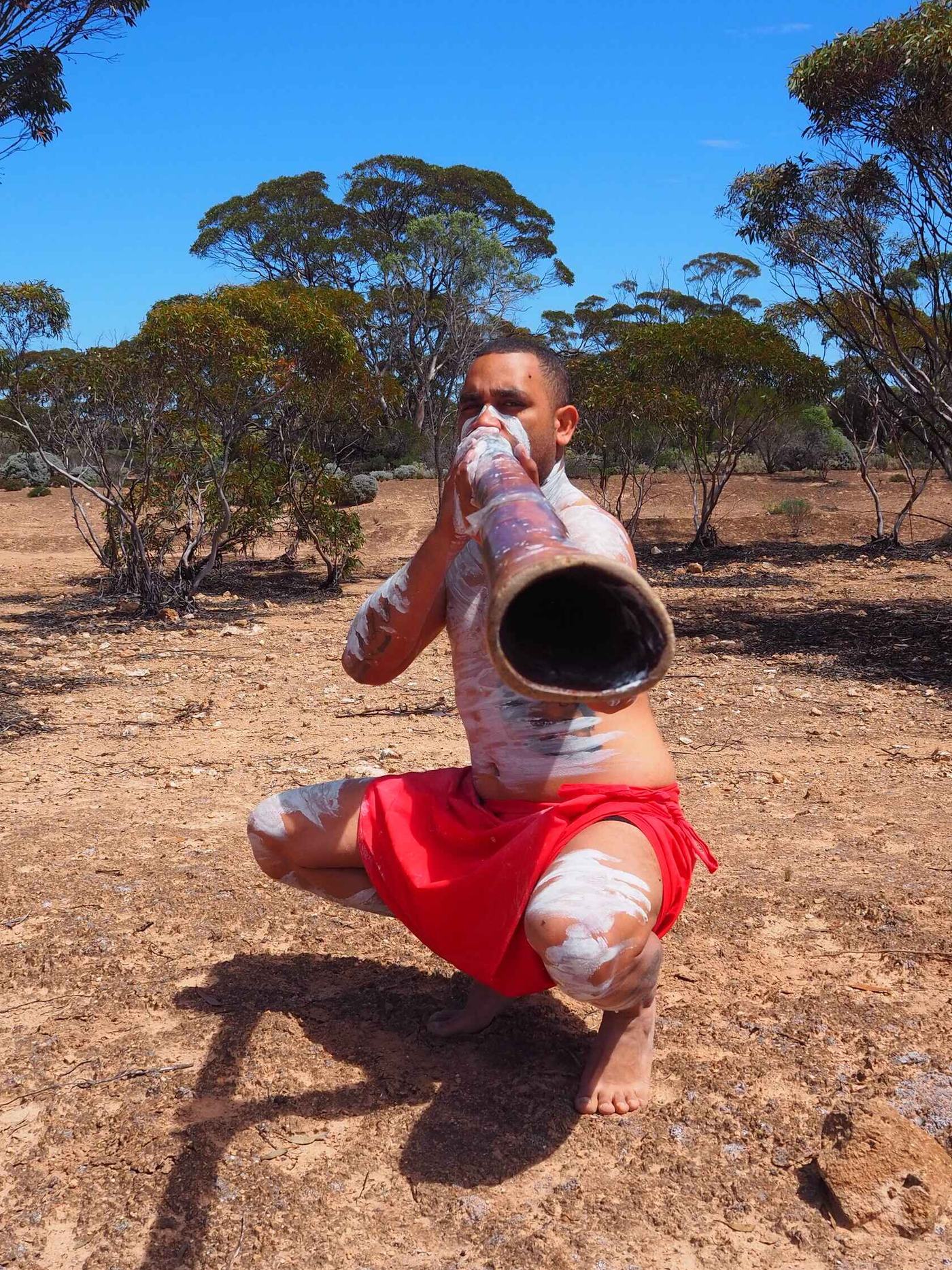Ryka Ali crouched on red earth playing the didgeridoo, mallee scrub behind him