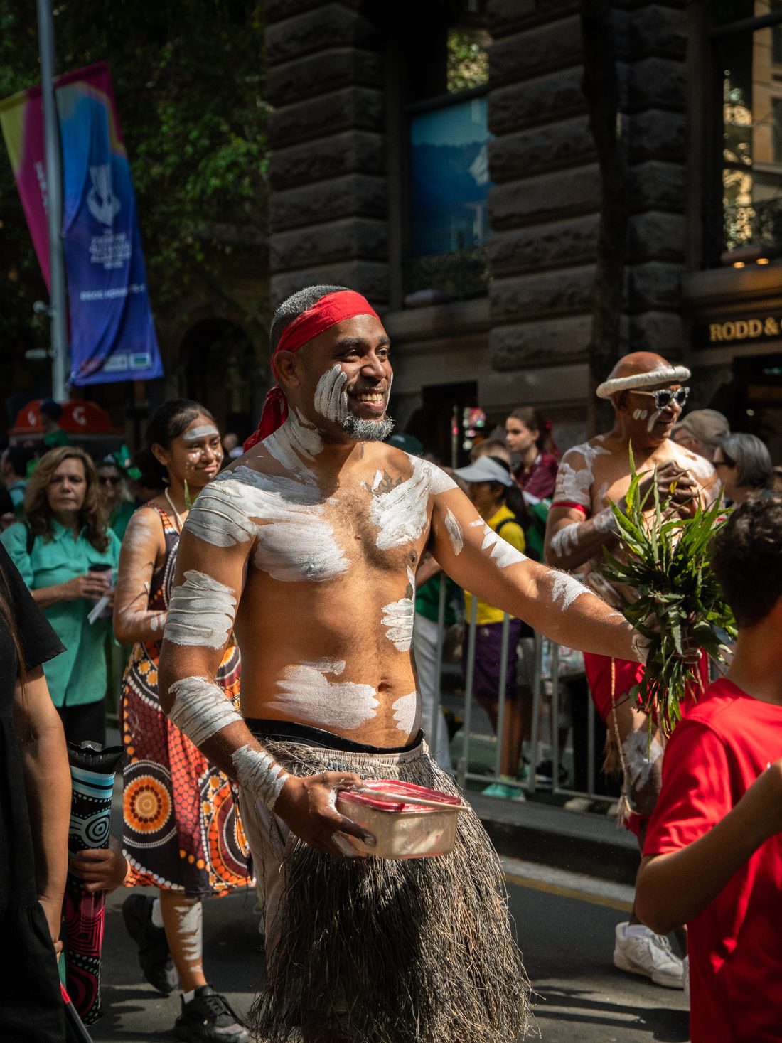 Ryka Ali smiling during a street procession, carrying smoking ceremony leaves